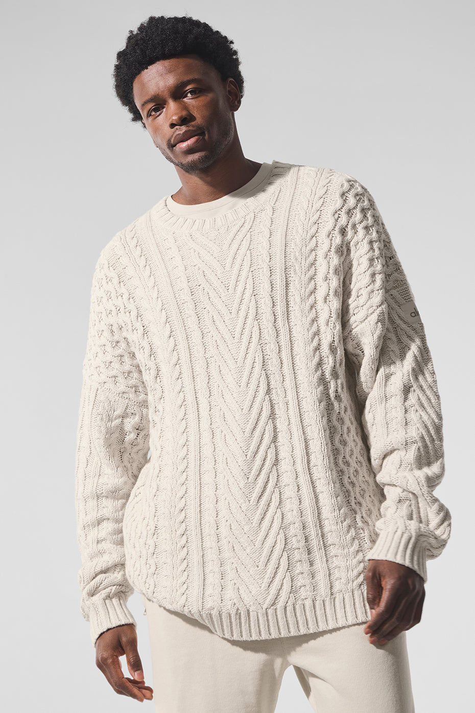 A man wearing a white cable knit sweater outdoors, layered over a denim shirt.