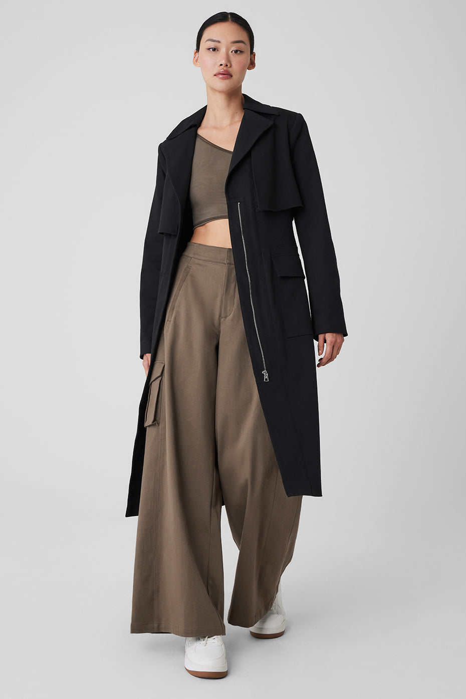 Model wearing a cropped trench coat with hands in pockets, urban backdrop.